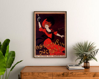 Vintage reklamaffisch Absinthe Ducros, inramad ovanför en träbyrå mellan gröna växter.