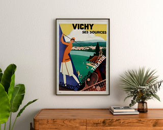 Vintage turistaffisch – Vichy, källor och turism, inramad ovanför en träbyrå, mellan växter.